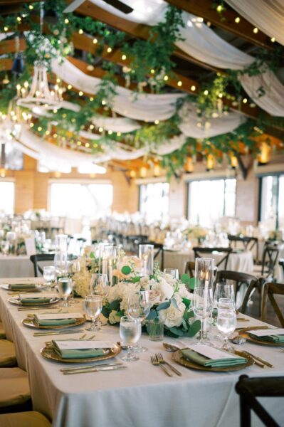 Elegant wedding reception with tables set with white linens, floral centerpieces, glassware, and candles. Greenery and draped fabric with twinkling lights hang from the wooden ceiling, creating a romantic atmosphere.