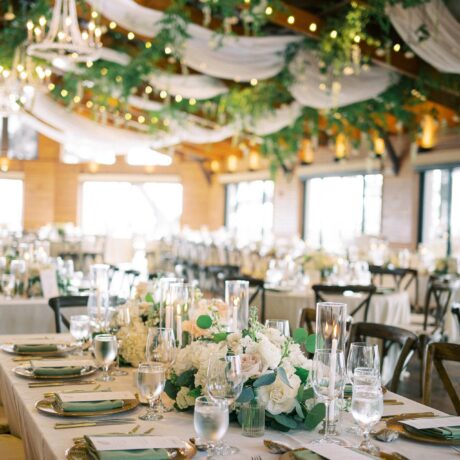 Elegant wedding reception with tables set with white linens, floral centerpieces, glassware, and candles. Greenery and draped fabric with twinkling lights hang from the wooden ceiling, creating a romantic atmosphere.