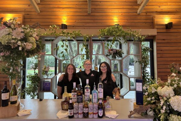 Three smiling bartenders in black outfits stand behind a decorated bar with various bottles of liquor and flowers, set in a wooden venue with large windows and greenery outside. Warm lighting creates a welcoming atmosphere.