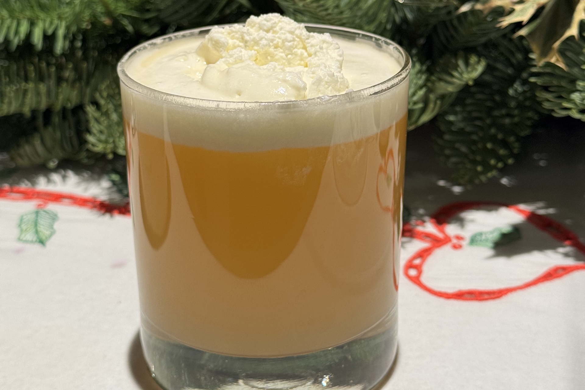 A glass of eggnog topped with whipped cream sits on a festive tablecloth, with green pine branches in the background.