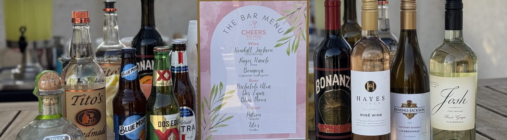 A selection of liquor and wine bottles arranged on a wooden table with a framed bar menu in the center; background shows an outdoor setting with trees and buildings.
