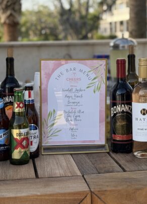 A selection of liquor and wine bottles arranged on a wooden table with a framed bar menu in the center; background shows an outdoor setting with trees and buildings.
