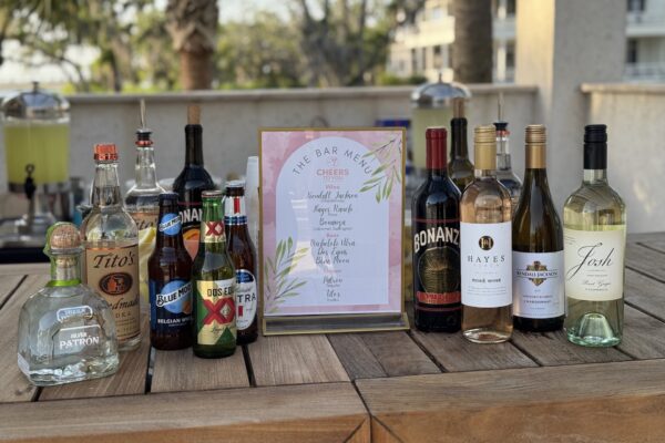 A selection of liquor and wine bottles arranged on a wooden table with a framed bar menu in the center; background shows an outdoor setting with trees and buildings.