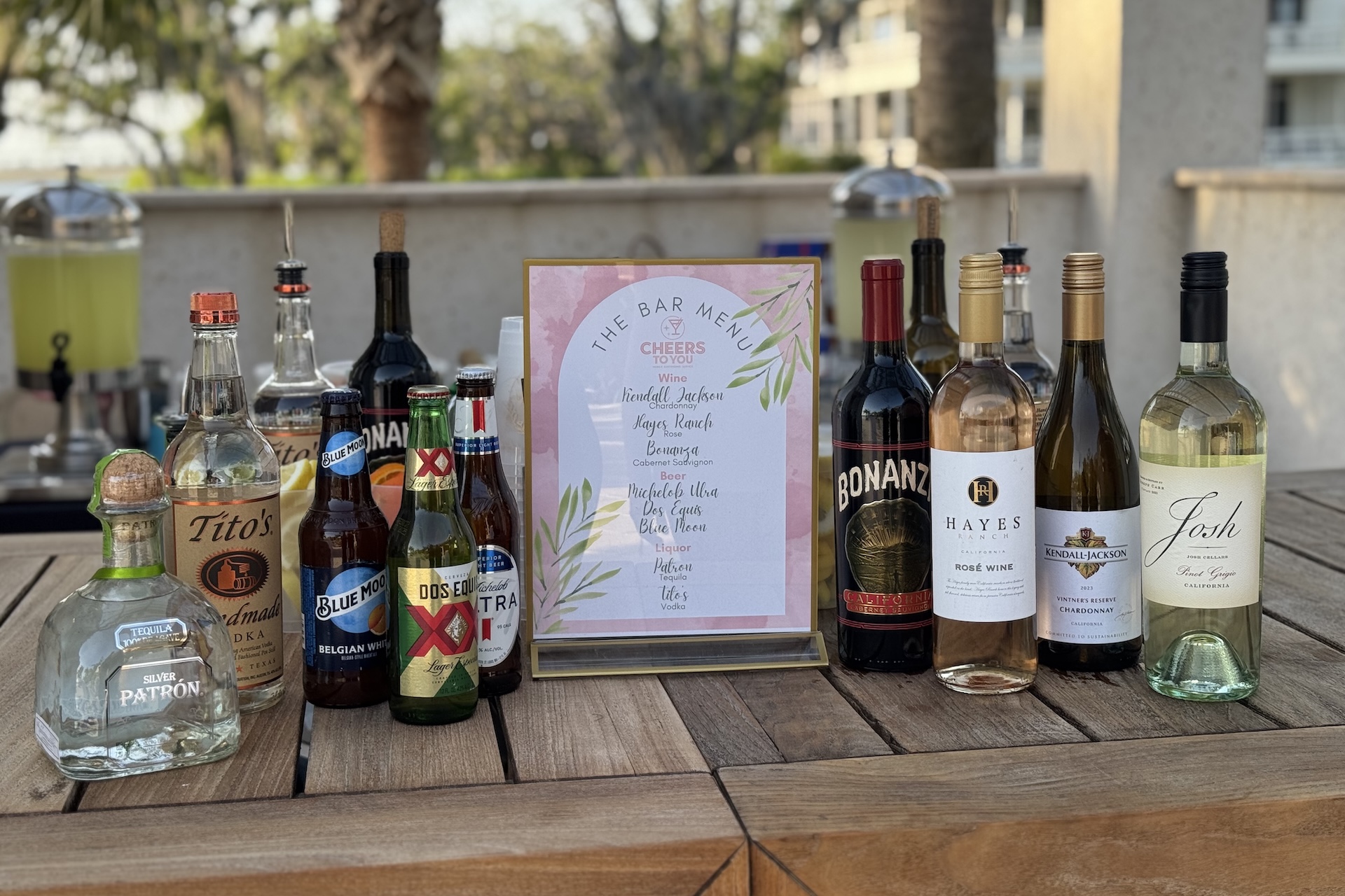 A selection of liquor and wine bottles arranged on a wooden table with a framed bar menu in the center; background shows an outdoor setting with trees and buildings.