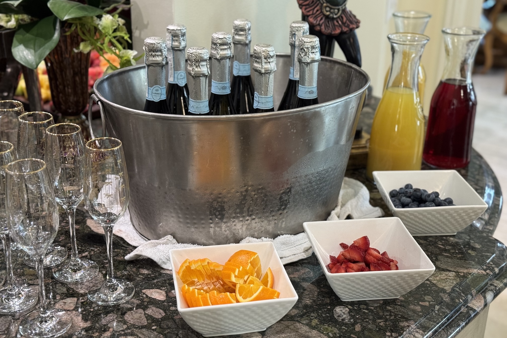 A large metal ice bucket holds bottles of sparkling wine, surrounded by empty champagne flutes, carafes of orange and red juice, and bowls of sliced oranges, strawberries, and blueberries on a countertop.