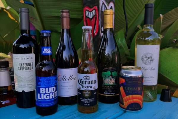 A selection of alcoholic beverages, including bottles of wine, beer, and a can, arranged on a blue table outdoors with green leaves and a colorful painted face in the background.