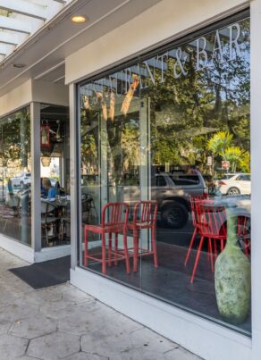 Street view of a modern restaurant with large windows, red chairs visible inside, a menu displayed outside, and trees reflecting on the glass facade. A brick sidewalk runs alongside the building.