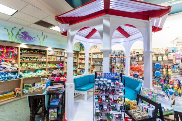 A colorful toy store with shelves full of stuffed animals, books, and various toys. A white gazebo with red trim and blue seats is in the center, and a display of die-cast toy cars is in the foreground.