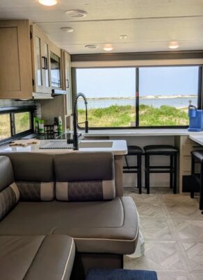 Interior of a modern RV with a brown sectional sofa, kitchenette, sink, large windows showing a scenic outdoor view of grass and water, barstools at the counter, and light wood cabinetry.