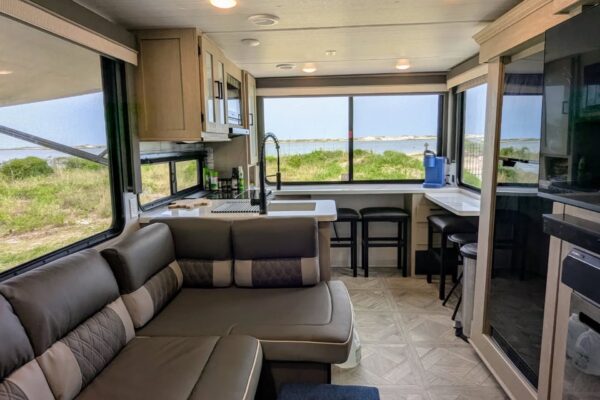 Interior of a modern RV with a brown sectional sofa, kitchenette, sink, large windows showing a scenic outdoor view of grass and water, barstools at the counter, and light wood cabinetry.