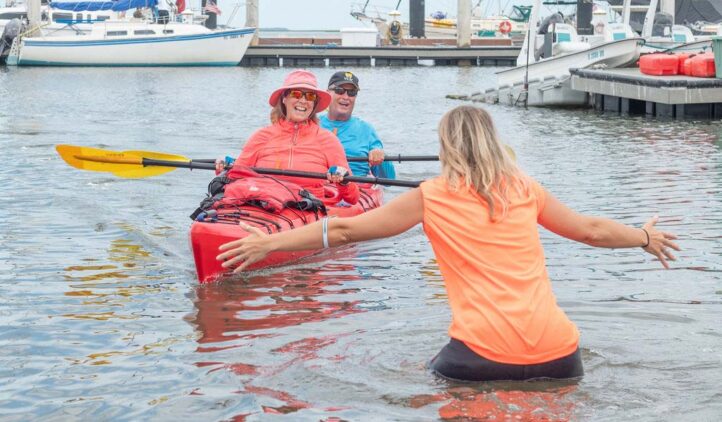 Two smiling people in a red kayak paddle toward a woman standing in shallow water with open arms, at a marina with several boats docked in the background.