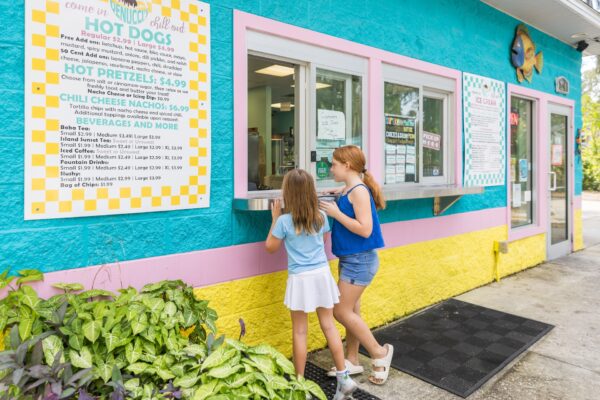 Two young girls stand at the window of a colorful outdoor food stand with a large menu on the wall, getting ready to order. The building is painted turquoise, yellow, and pink.