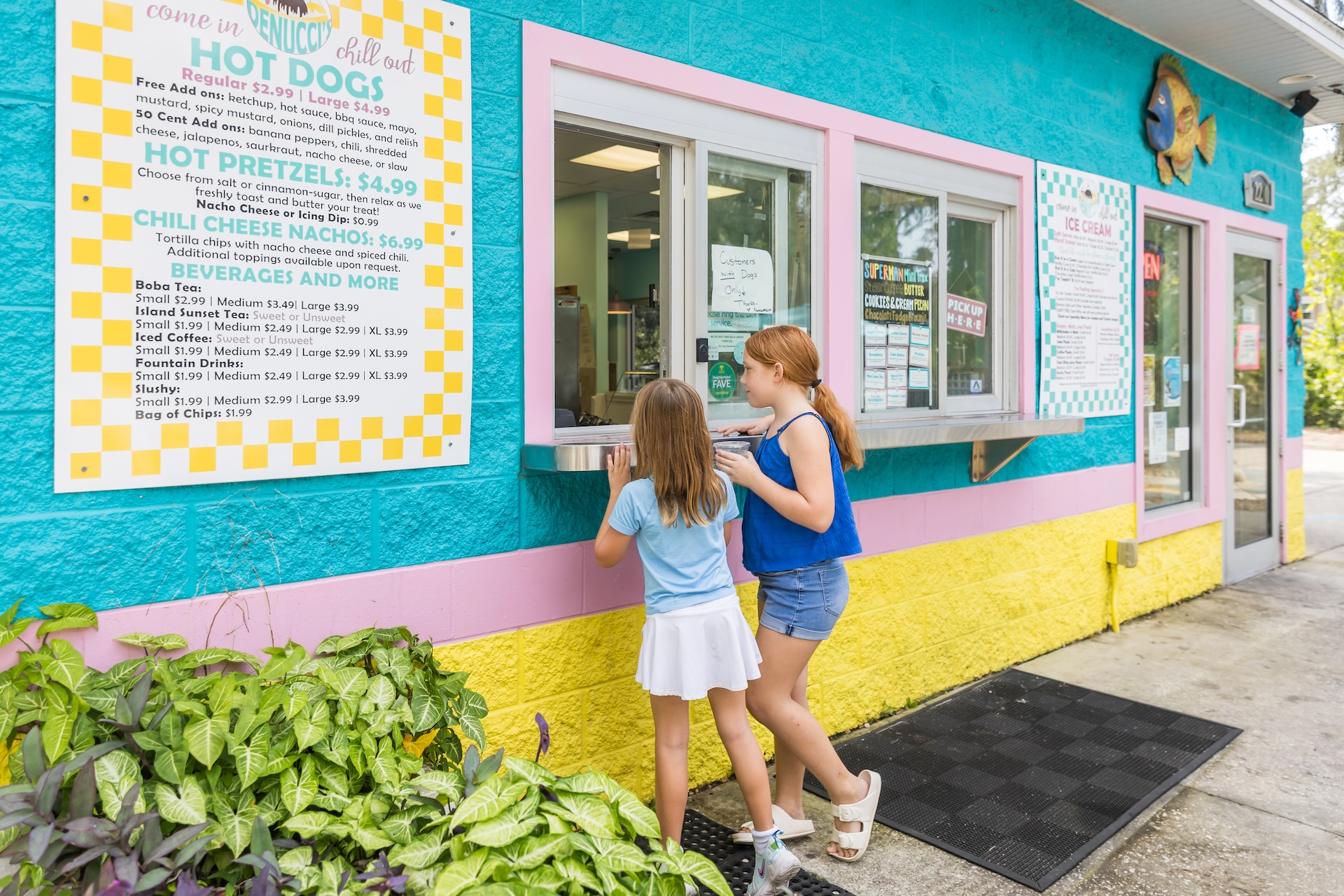 Two young girls stand at the window of a colorful outdoor food stand with a large menu on the wall, getting ready to order. The building is painted turquoise, yellow, and pink.