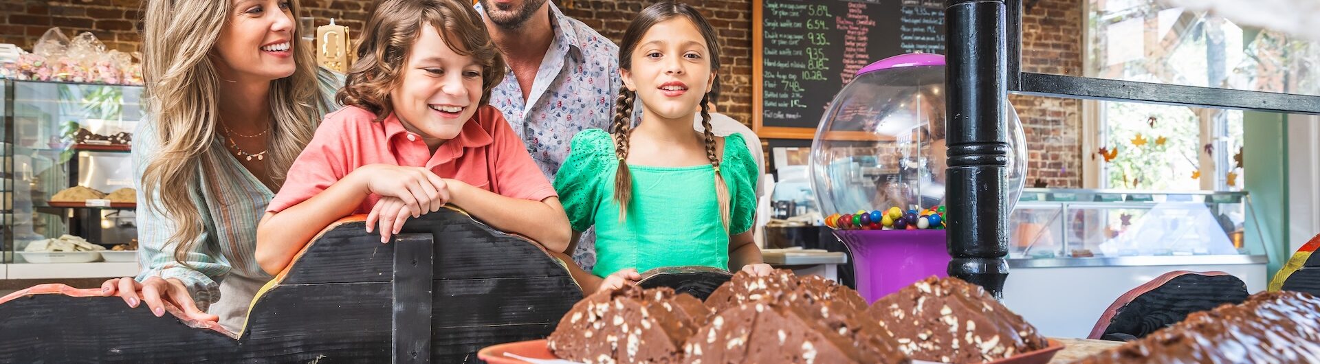 A smiling family of four admires a display of large chocolate treats inside a bakery or candy shop, with a chalkboard menu and gumball machine visible in the background.
