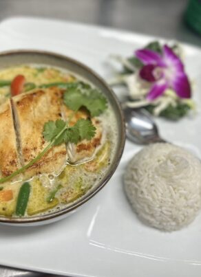 A bowl of Thai green curry with grilled chicken, vegetables, and cilantro garnish, served with a mound of white rice and a small side salad with an edible purple orchid on a white plate. A spoon is placed beside the rice.