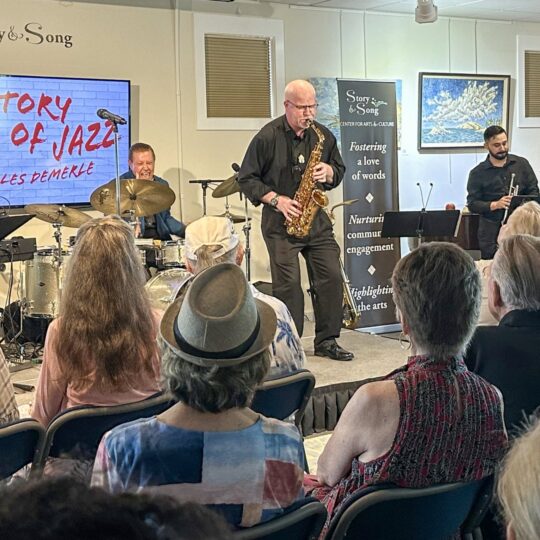 A jazz band performs on stage for an audience. A screen beside them reads The Story of Jazz with Les DeMerle. People sit attentively, some wearing hats. Musical instruments and art decorate the well-lit venue.
