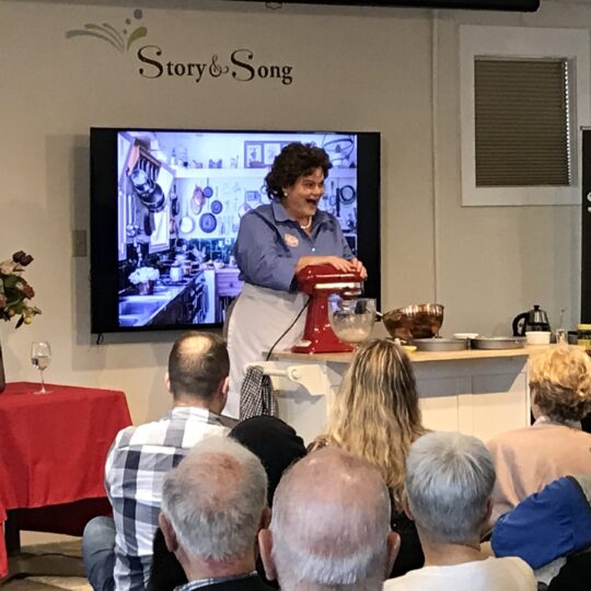 A woman in costume demonstrates cooking with a mixer on stage at the Story & Song Center for Arts & Culture, addressing an audience. A photo of a kitchen is displayed on a screen behind her.