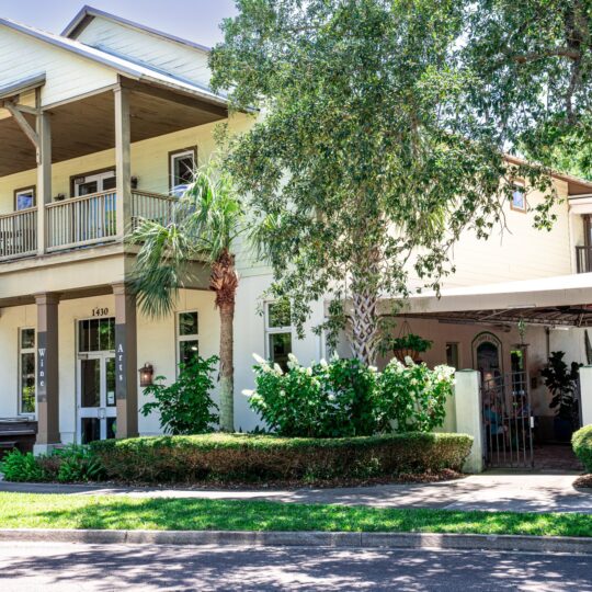 A two-story light yellow building with a wraparound balcony, surrounded by green trees and bushes. A covered outdoor seating area and sign reading Sugarbakers Bistro are visible near the entrance.