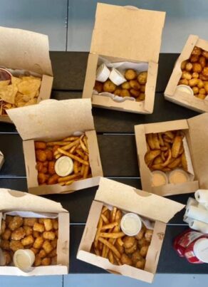 An overhead view of a table with several open takeout boxes filled with fries, tater tots, chips, curly fries, dipping sauces, and two stacks of napkins with packets of ketchup.