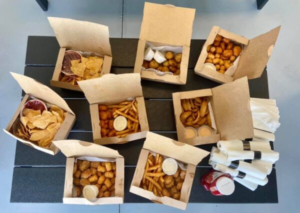 An overhead view of a table with several open takeout boxes filled with fries, tater tots, chips, curly fries, dipping sauces, and two stacks of napkins with packets of ketchup.