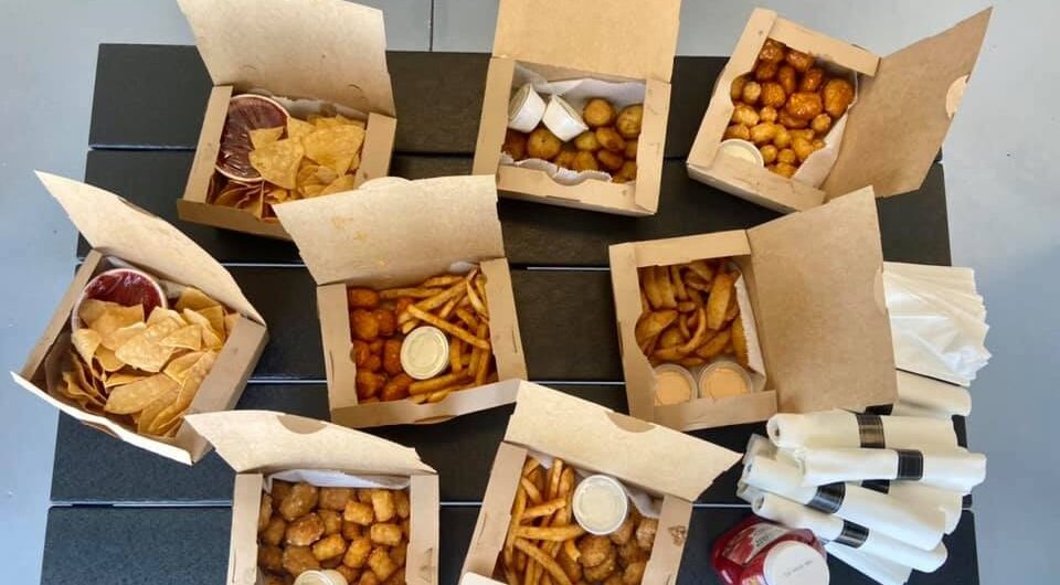An overhead view of a table with several open takeout boxes filled with fries, tater tots, chips, curly fries, dipping sauces, and two stacks of napkins with packets of ketchup.