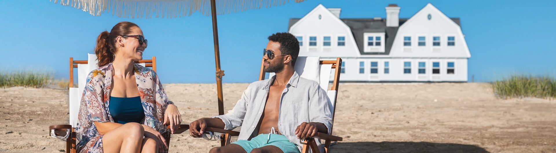 A woman and a man relax on beach chairs under a large white umbrella, smiling at each other. A white house with blue shutters sits in the background on a sunny day.