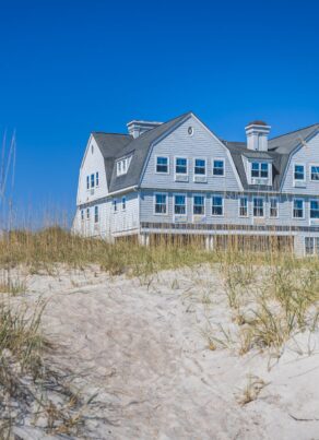 A large, gray beach house with many windows sits behind dunes covered in tall grass, under a clear blue sky.