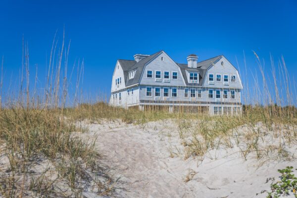 A large, gray beach house with many windows sits behind dunes covered in tall grass, under a clear blue sky.