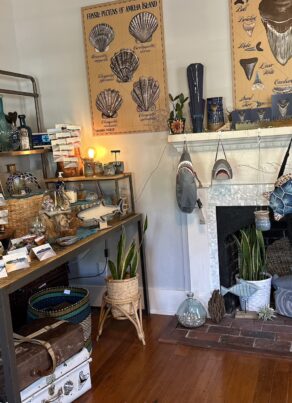 A cozy shop corner displays jewelry, pottery, baskets, and sea-themed decor. Necklaces hang on stands and boards; wall posters feature shells and whale anatomy above a white fireplace filled with plants and nautical items.