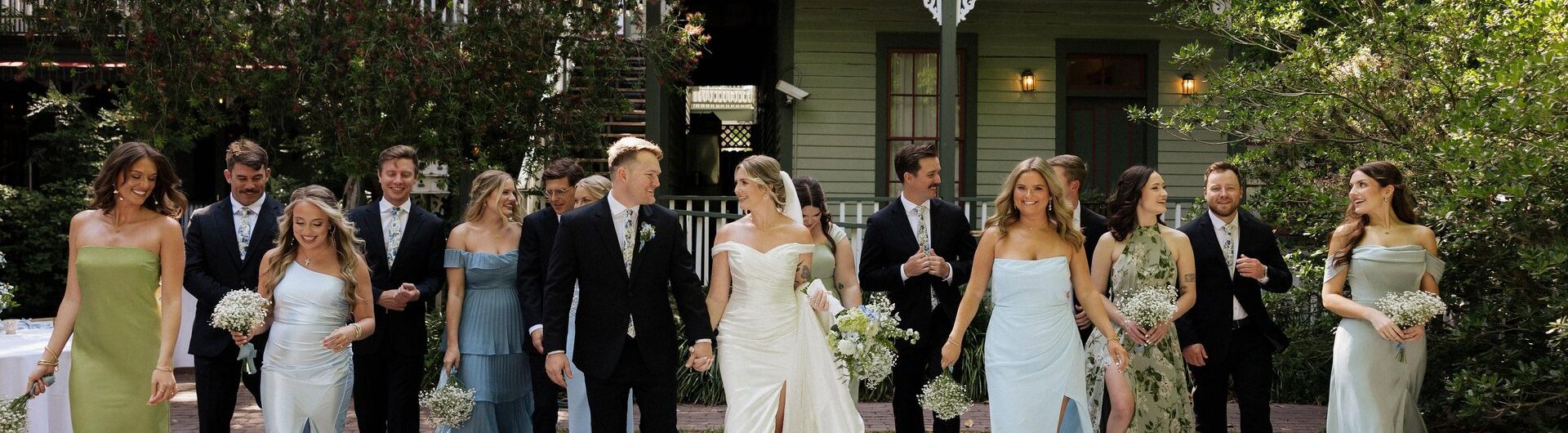 A wedding party poses outdoors in front of a green house, with bridesmaids in pastel dresses and groomsmen in dark suits. The bride and groom stand in the center, holding hands, on a large black-and-white checkered floor.