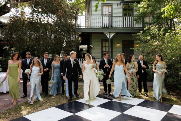 A wedding party poses outdoors in front of a green house, with bridesmaids in pastel dresses and groomsmen in dark suits. The bride and groom stand in the center, holding hands, on a large black-and-white checkered floor.