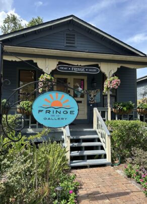 A small blue house with a porch, plants, and hanging flowers. A large round sign in front reads “Fringe Art Gallery.” A whimsical dog sculpture is visible near the entrance.