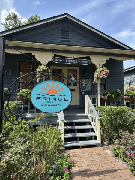 A small blue house with a porch, plants, and hanging flowers. A large round sign in front reads “Fringe Art Gallery.” A whimsical dog sculpture is visible near the entrance.