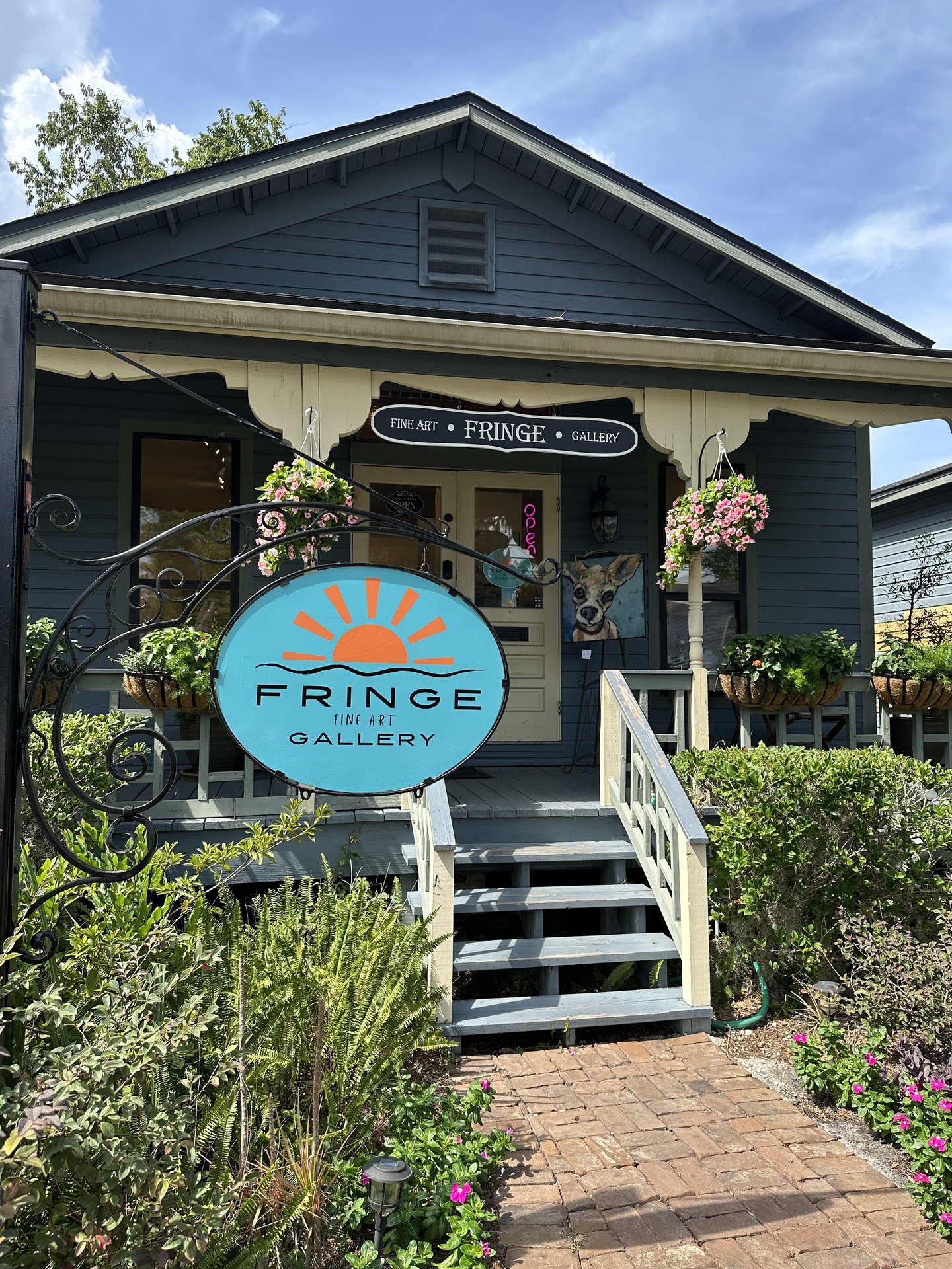 A small blue house with a porch, plants, and hanging flowers. A large round sign in front reads “Fringe Art Gallery.” A whimsical dog sculpture is visible near the entrance.