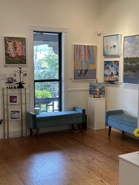 A bright art gallery room with wooden floors, light walls, blue benches, and colorful paintings. Artwork includes figures, beach scenes, and landscapes. A window lets in natural light, with greenery visible outside.