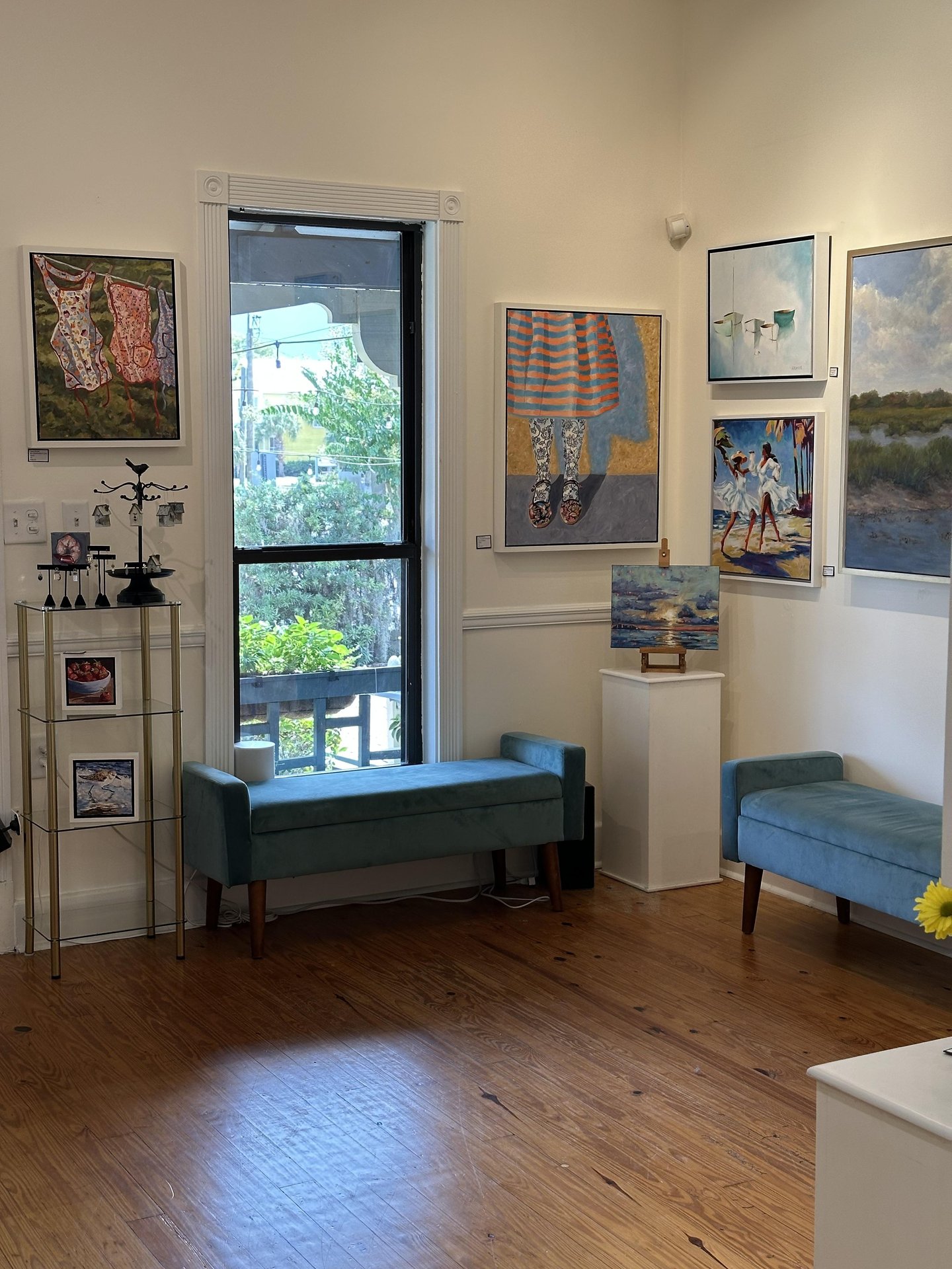 A bright art gallery room with wooden floors, light walls, blue benches, and colorful paintings. Artwork includes figures, beach scenes, and landscapes. A window lets in natural light, with greenery visible outside.