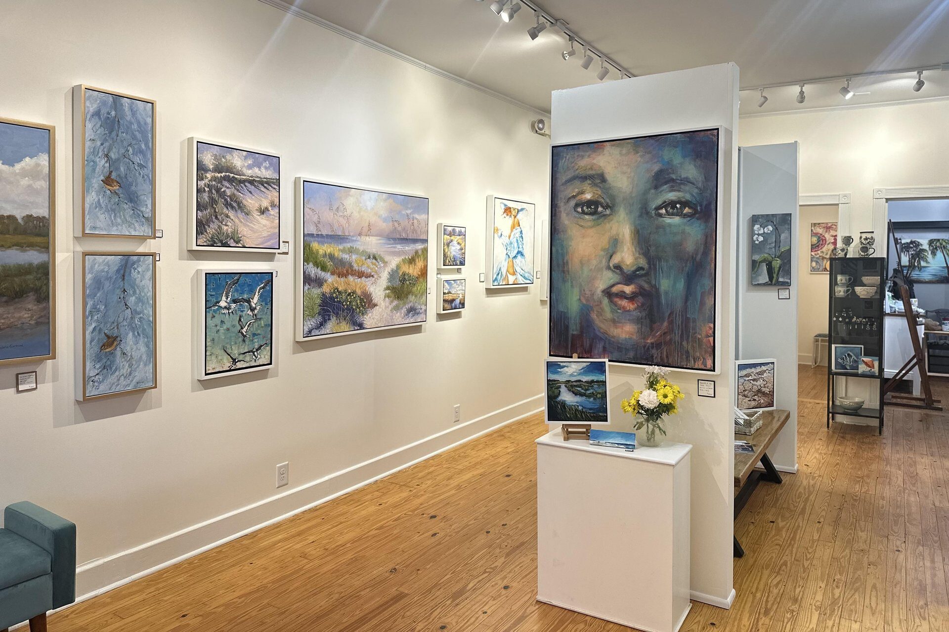 An art gallery with wooden floors and white walls displays various landscape paintings and a large portrait of a face. There is a small table with flowers and brochures, and artwork arranged on the surrounding walls and shelves.