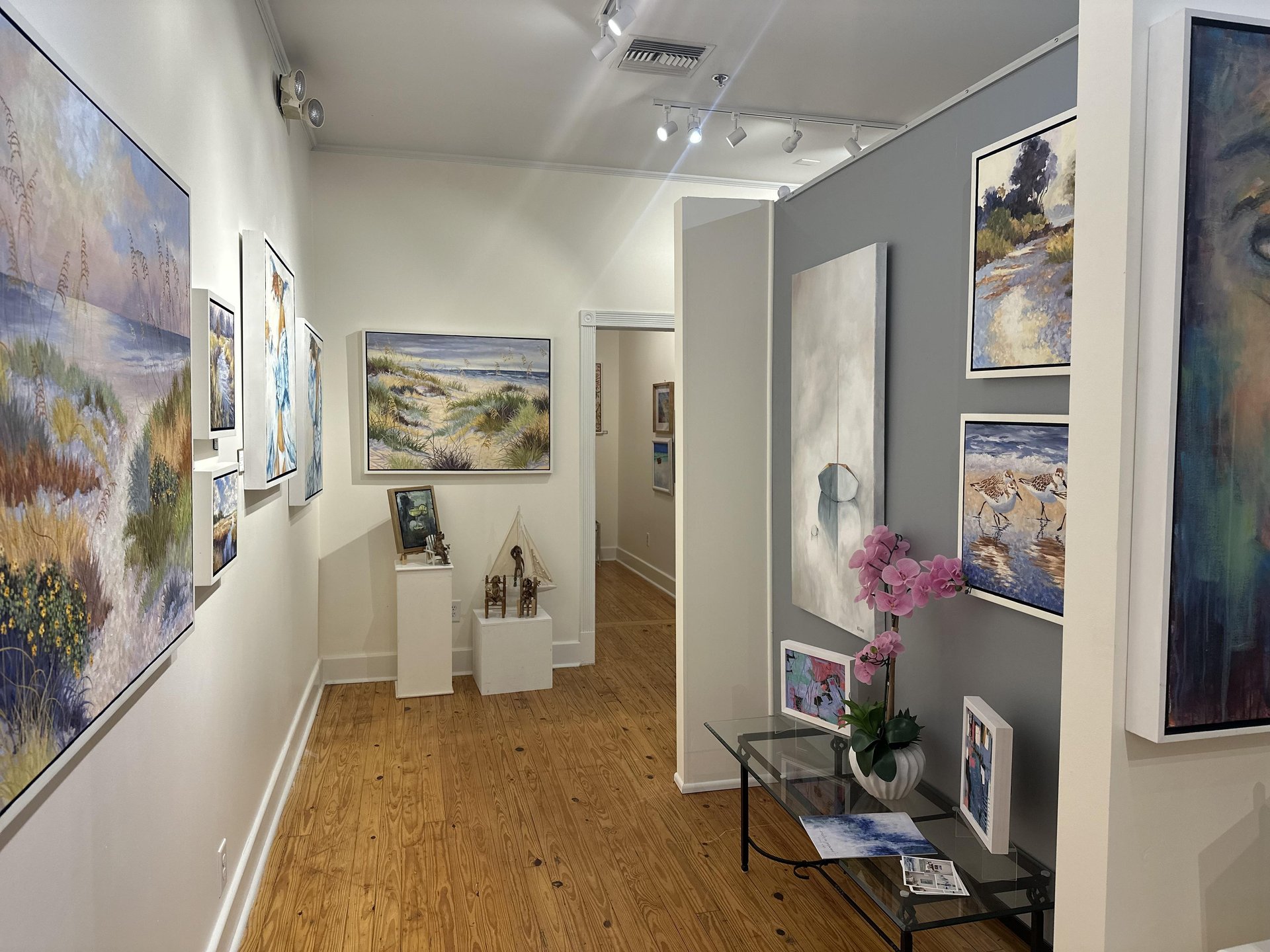 An art gallery with landscape paintings on white walls, wood flooring, a glass table with a pink orchid and books, and sculptures on a pedestal. The space is well-lit, with a hallway leading further into the gallery.