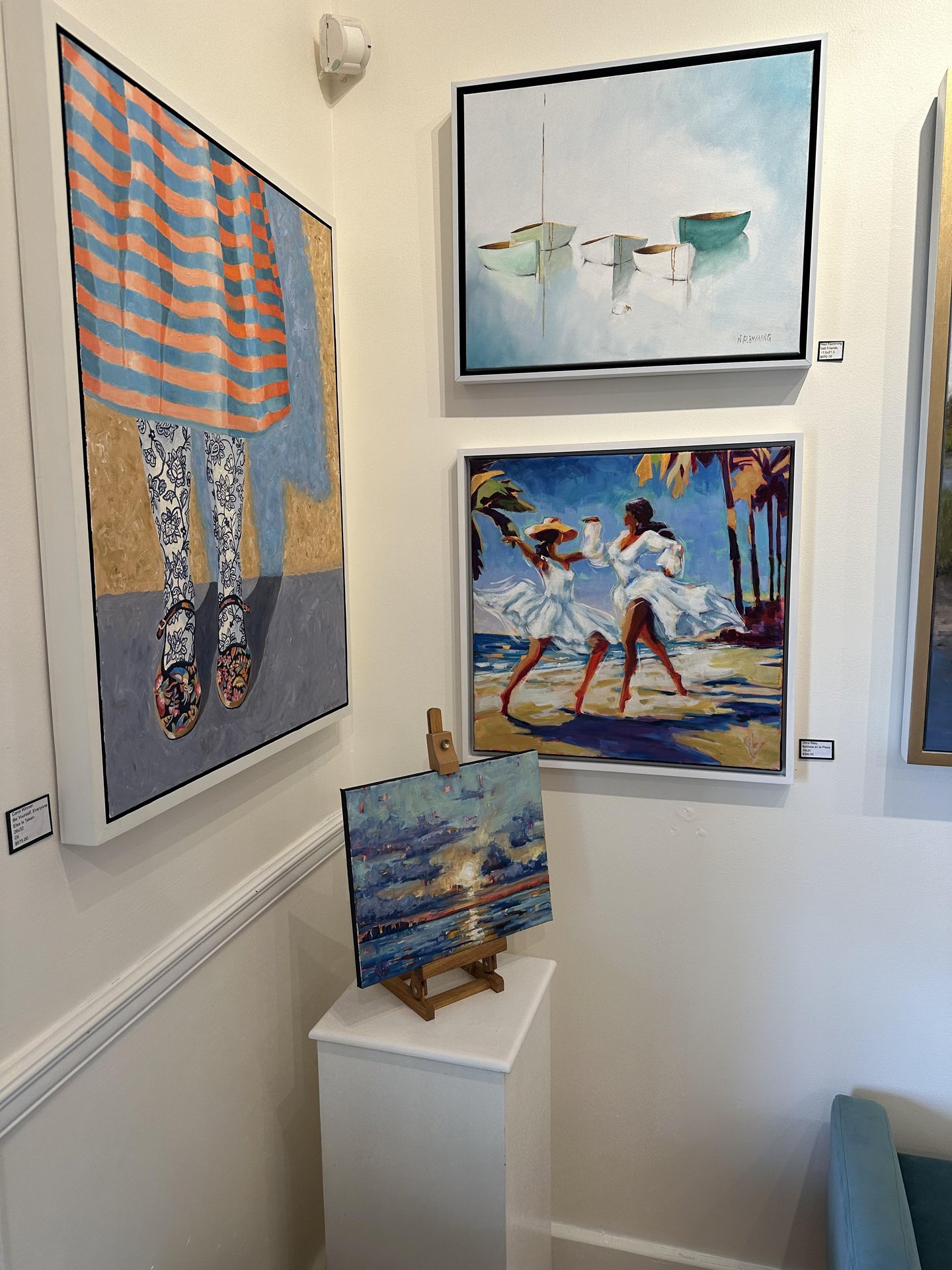 A gallery wall displaying four colorful paintings: a figure in a striped skirt, boats on water, two dancers on a beach, and a small seascape on an easel atop a white pedestal.