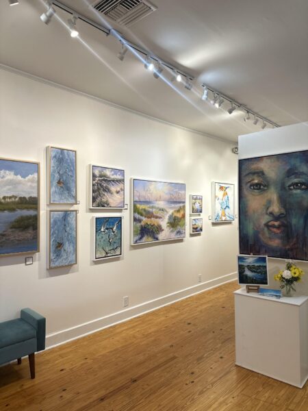 A bright art gallery with landscape paintings on a white wall, a large portrait of a face on the right, a small bench, and a display pedestal with flowers and brochures on a wooden floor.
