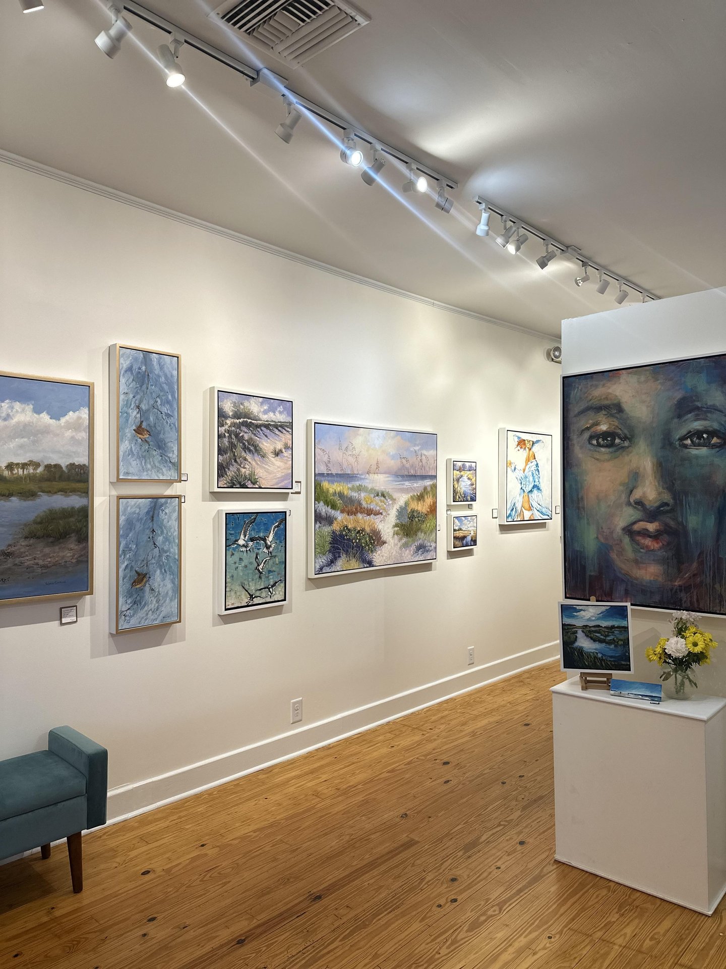 A bright art gallery with landscape paintings on a white wall, a large portrait of a face on the right, a small bench, and a display pedestal with flowers and brochures on a wooden floor.