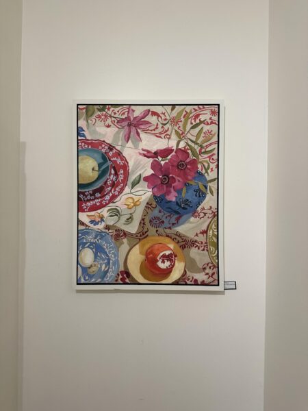 A colorful painting hangs on a white wall, depicting a vase of vibrant flowers, patterned dishes, fruit, and a red-and-white decorative tablecloth with intricate floral designs.