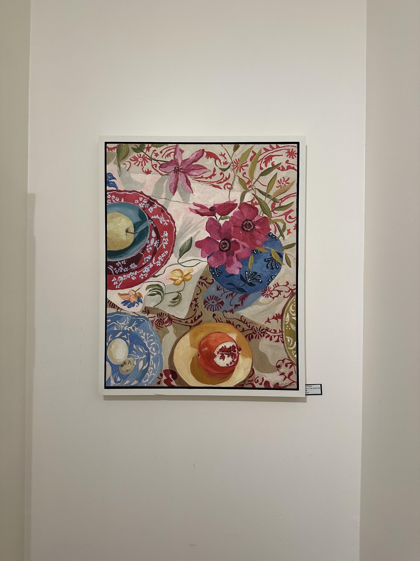 A colorful painting hangs on a white wall, depicting a vase of vibrant flowers, patterned dishes, fruit, and a red-and-white decorative tablecloth with intricate floral designs.