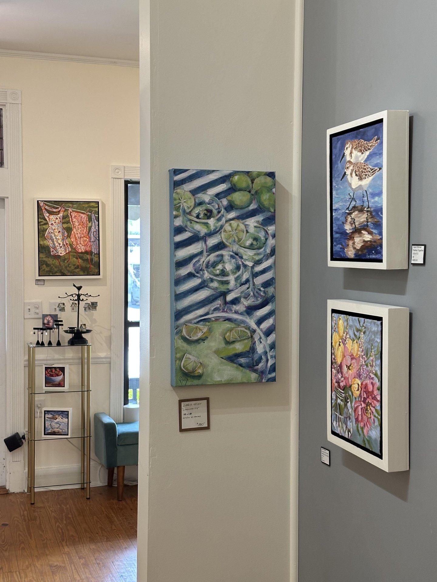 A small art gallery with white walls displays colorful paintings, including cocktails, a bird, flowers, and dresses. A metal sculpture sits on a shelf, and a teal chair is visible near a sunlit window.