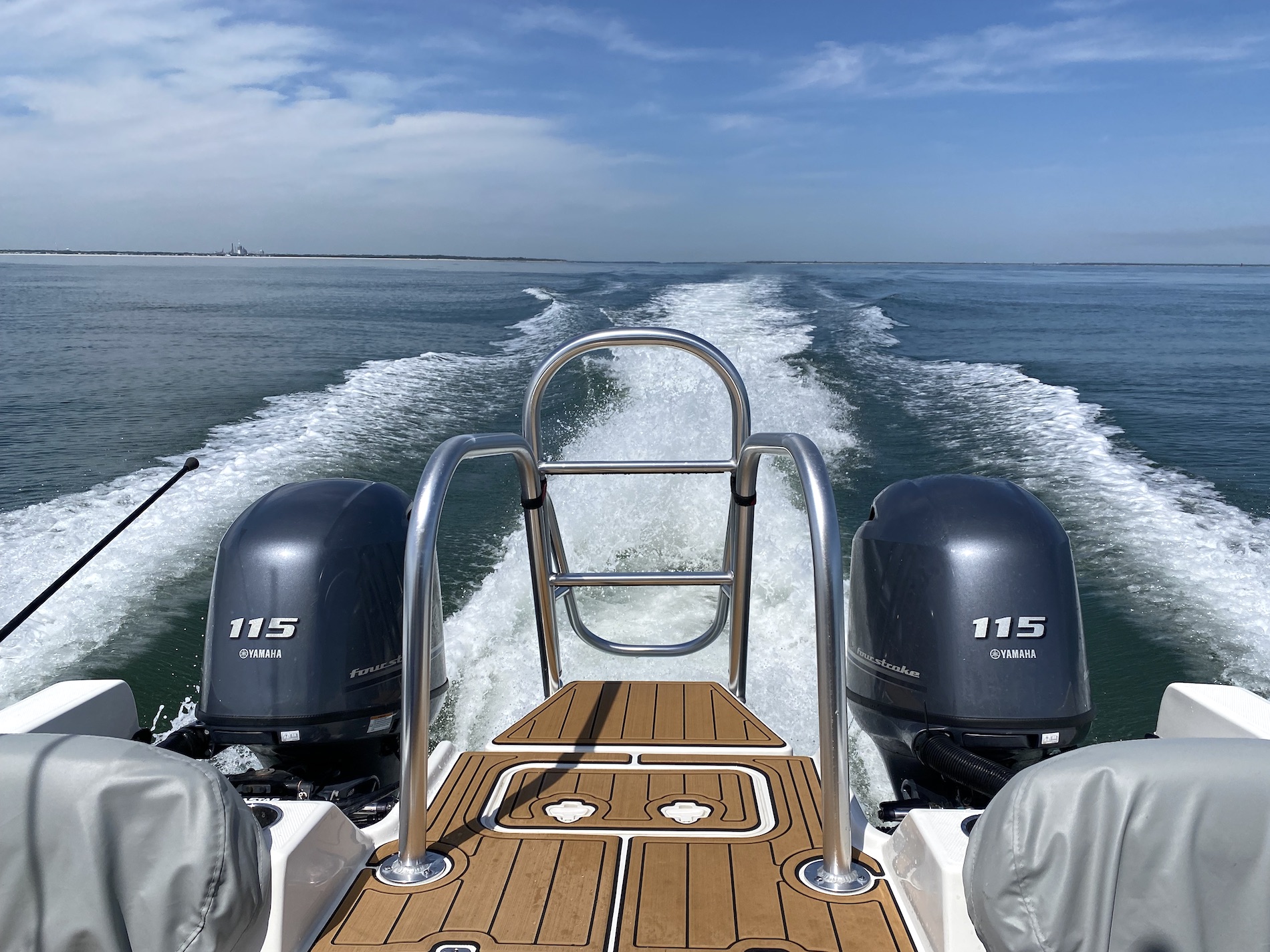 View from the back of a boat with two Yamaha 115 outboard motors, wooden deck, and metal railings, moving quickly on calm blue water, leaving a trail of white wake under a partly cloudy sky.