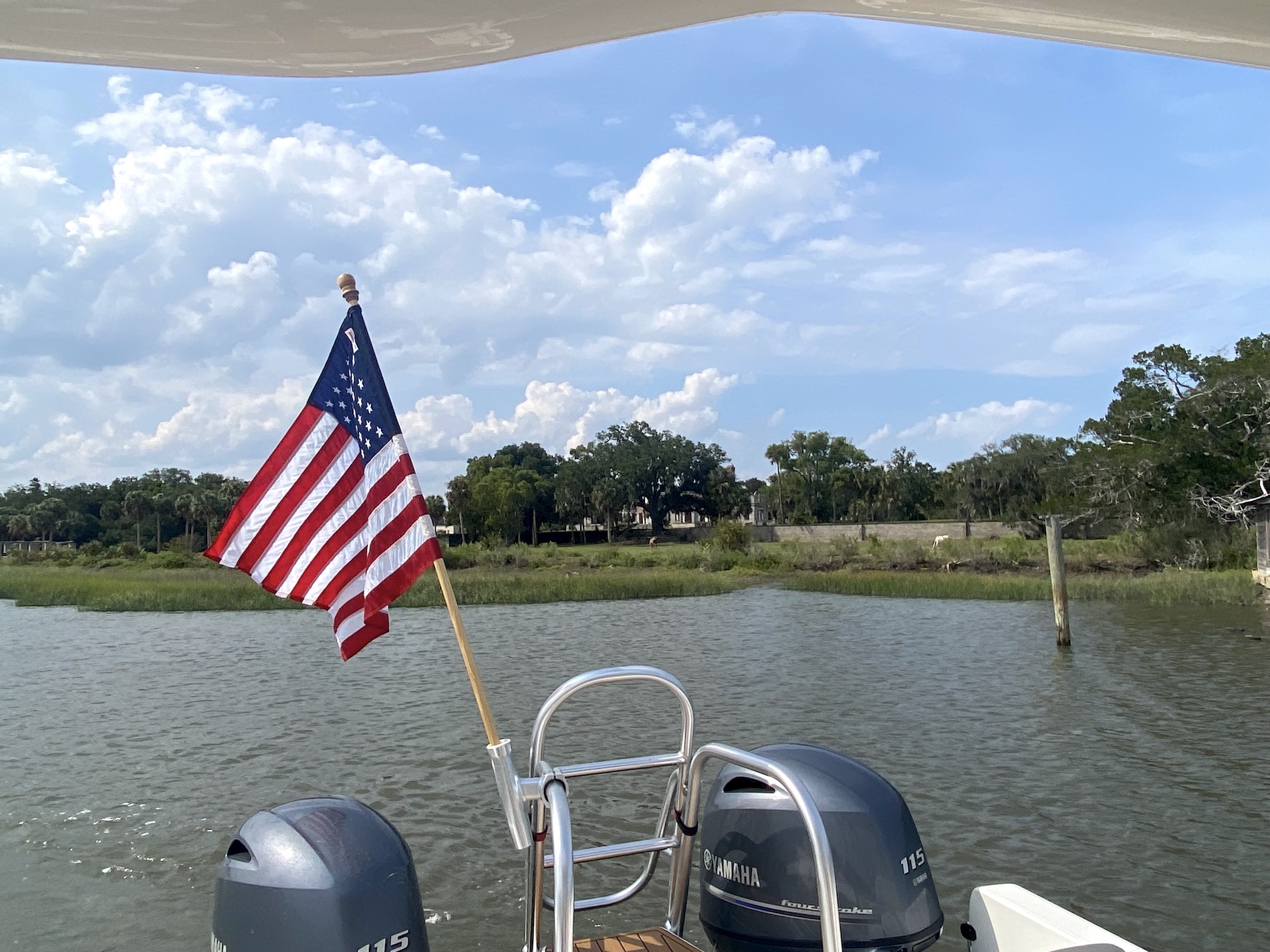 A boat with two Yamaha motors and an American flag is on calm water near a grassy shoreline with trees under a blue sky with scattered clouds.