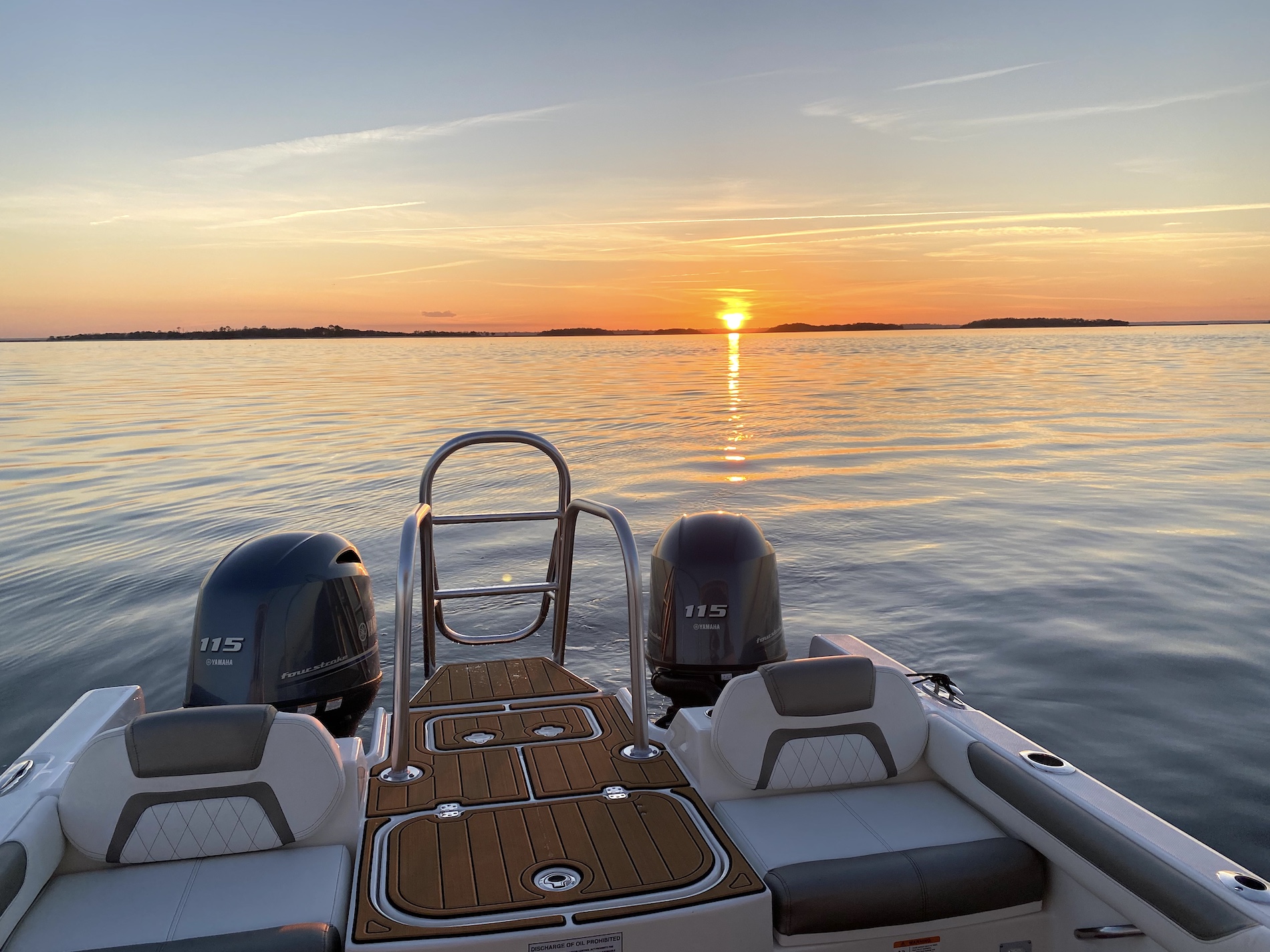 A view from the back of a boat with two outboard motors, floating on calm water at sunset; the sun is low on the horizon, casting a golden reflection over the water.
