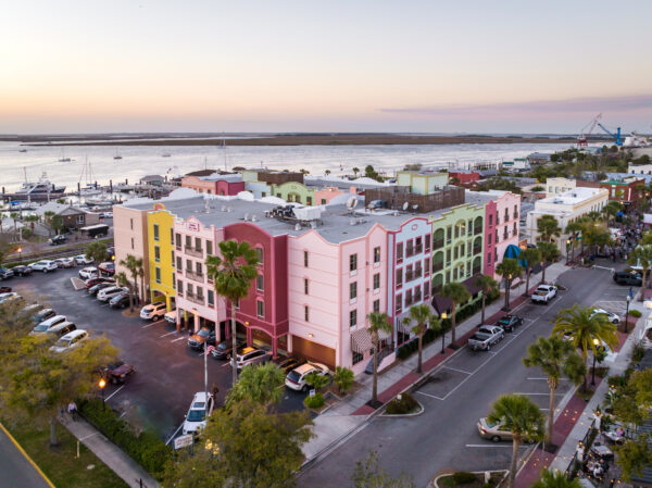 Aerial view of pastel-colored buildings and palm trees along a street near a marina at sunset, with cars parked and boats docked by the waterfront.