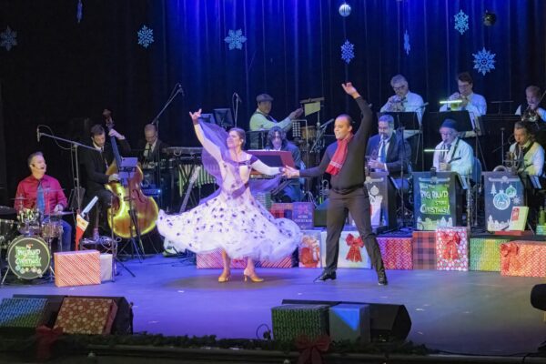 A woman in a flowing white dress and a man in a black outfit with a red scarf dance on stage, surrounded by Christmas decorations and gifts. A live band plays in the background under blue stage lights and hanging snowflakes.