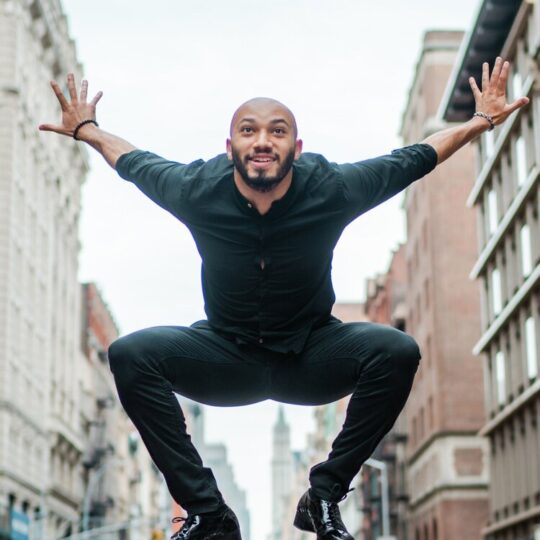 A man in a black outfit jumps mid-air on a city street, arms spread wide and smiling, with tall buildings lining both sides of the street and cars visible in the background.